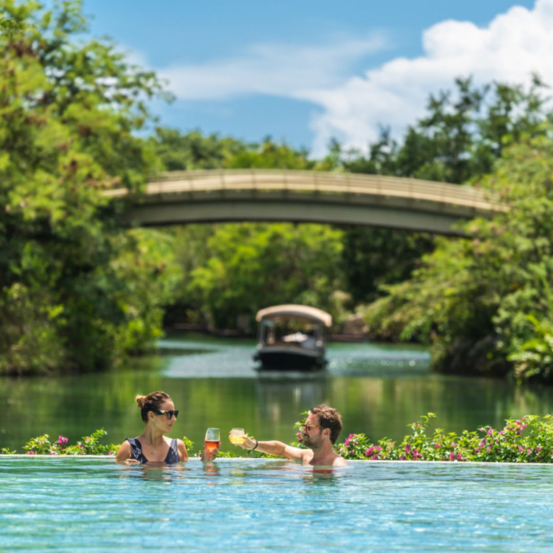 Man and woman in sunglasses cheering cocktails in a pool with a boat floating on green lagoon, a bridge and lots of lush greenery in the background.
