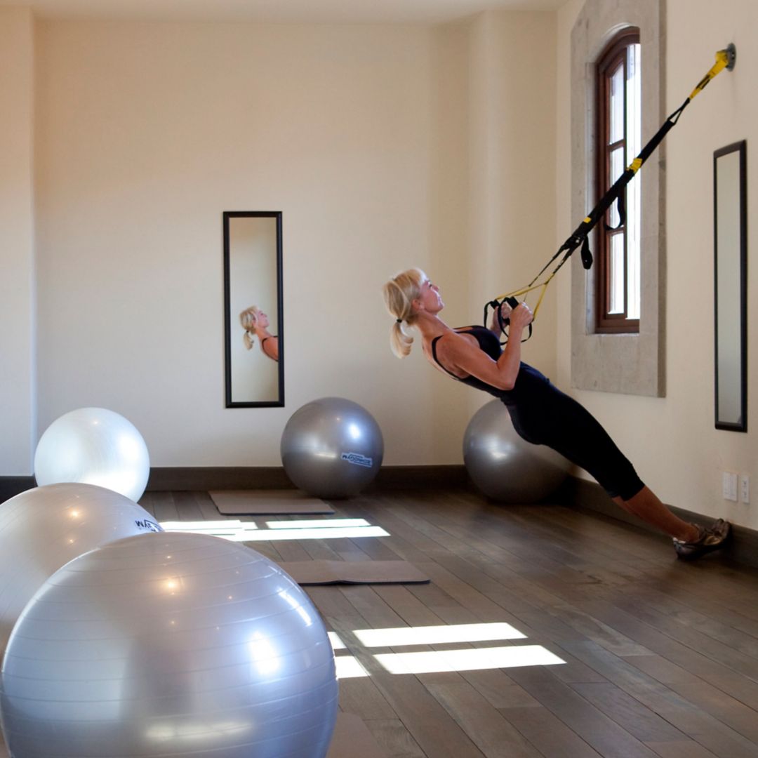A woman performing a TRX suspension exercise in a room with exercise balls and yoga mats.