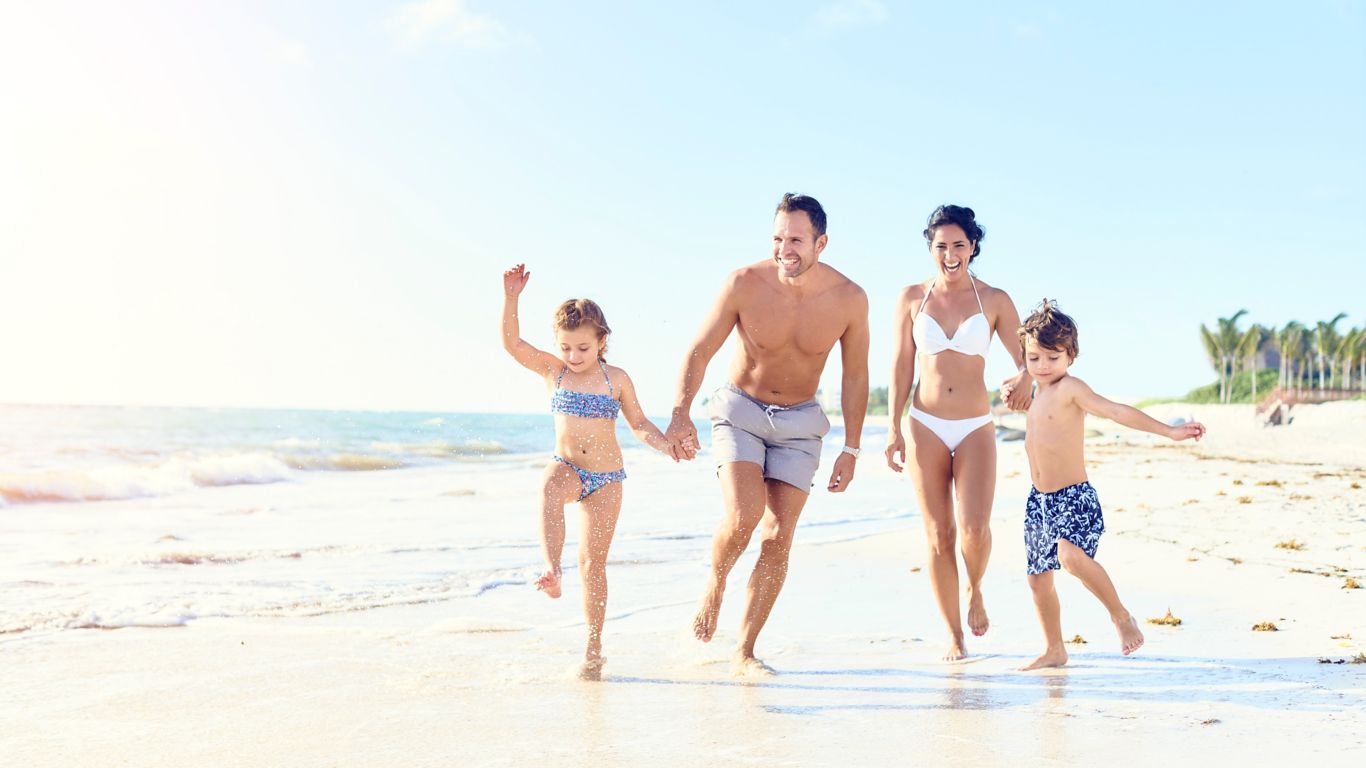 pais e duas crianças pequenas (menino e menina) de mãos dadas enquanto correm pela areia perto da água em trajes de banho