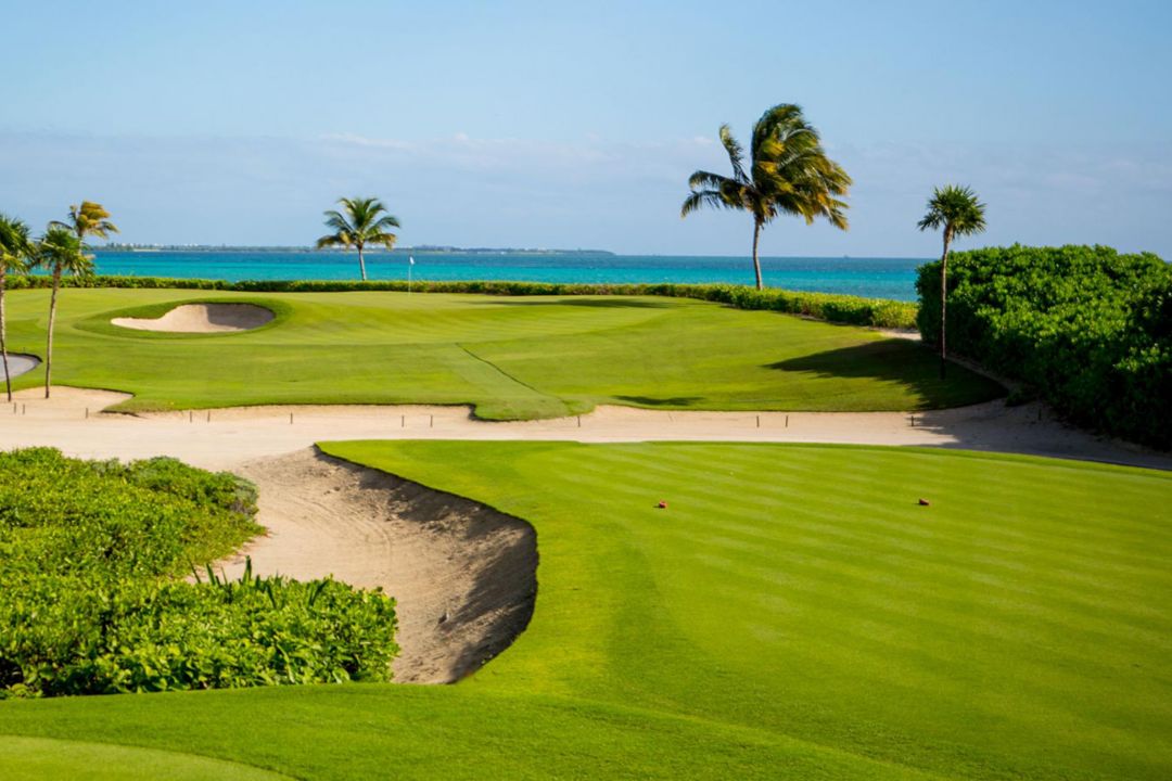 Vista de los campos de golf del campeonato El Camaleón de color verde brillante, pozos de arena, con palmeras y aguas turquesas de fondo
