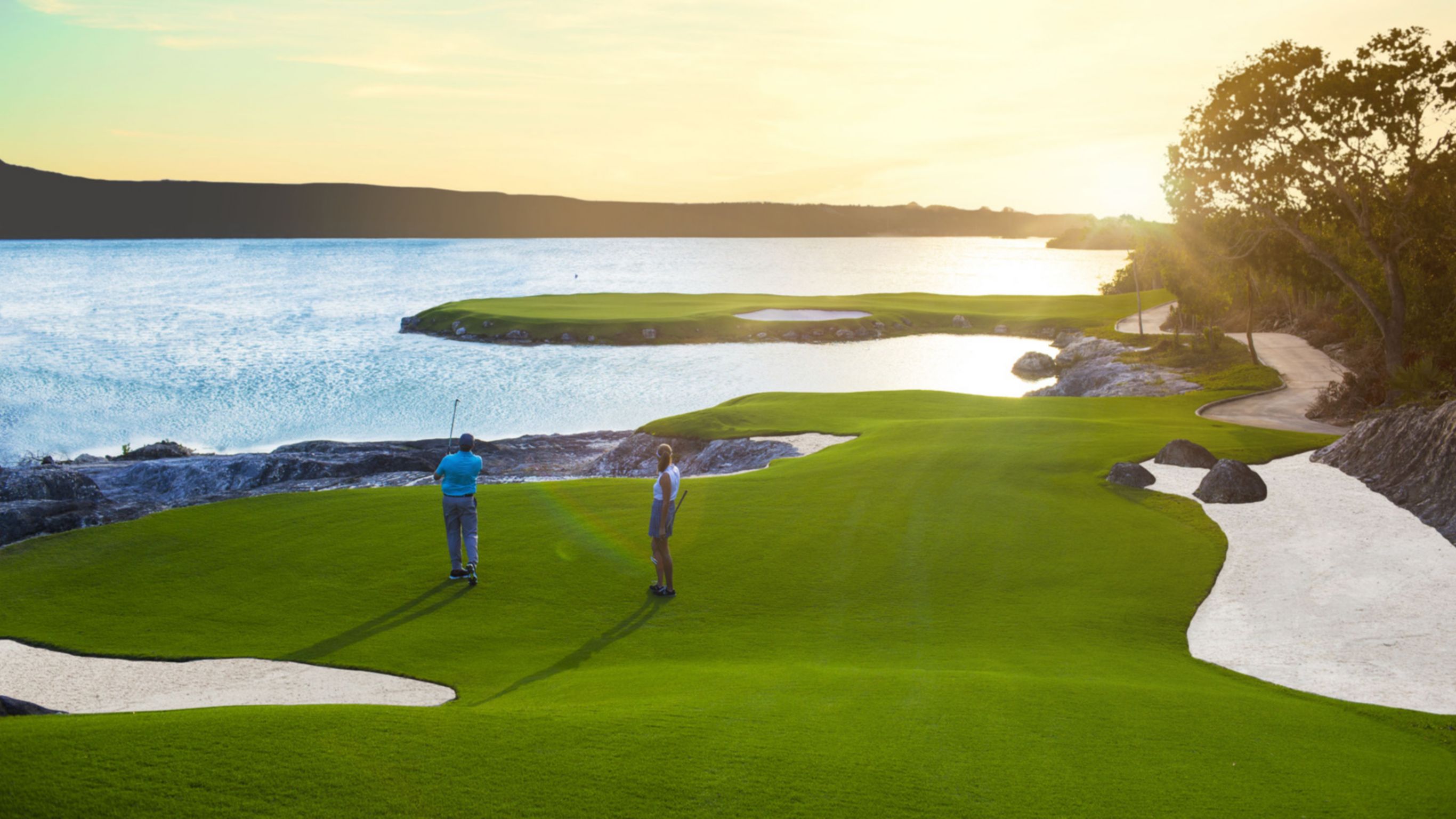 Two people playing golf on a green near a body of water, with the sun setting in the background.