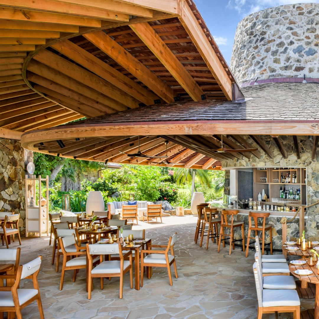 Beachside hotel bar with square tables arranged and a round mirror on the right side stone wall.