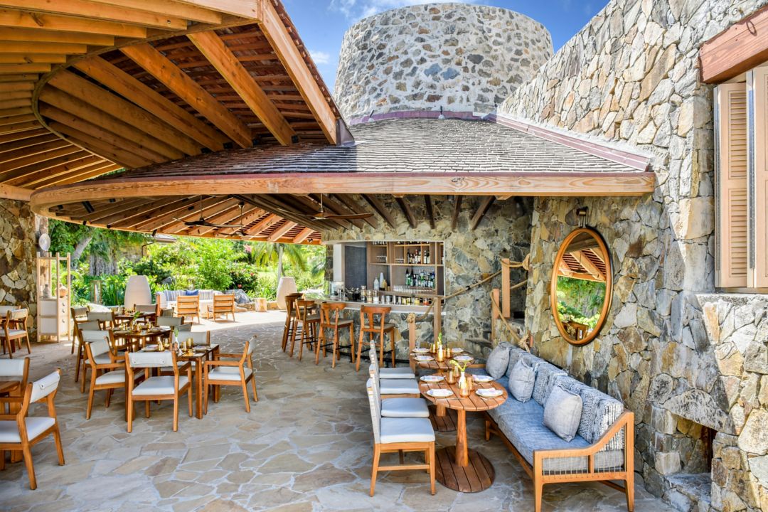 Beachside hotel bar with square tables arranged and a round mirror on the right side stone wall.