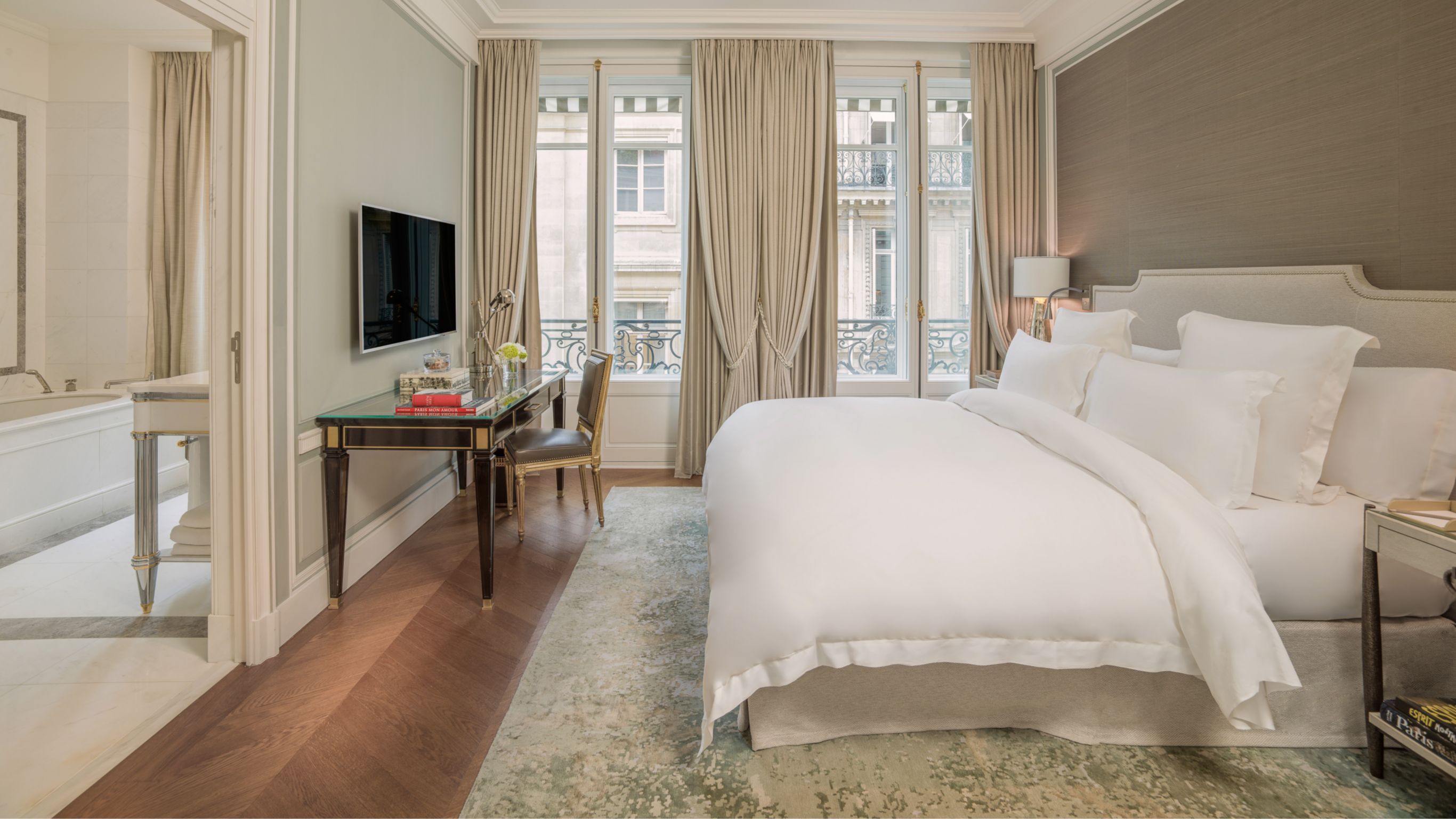 A Parisian hotel suite with a king bed, white linens, large windows, and beige curtains.