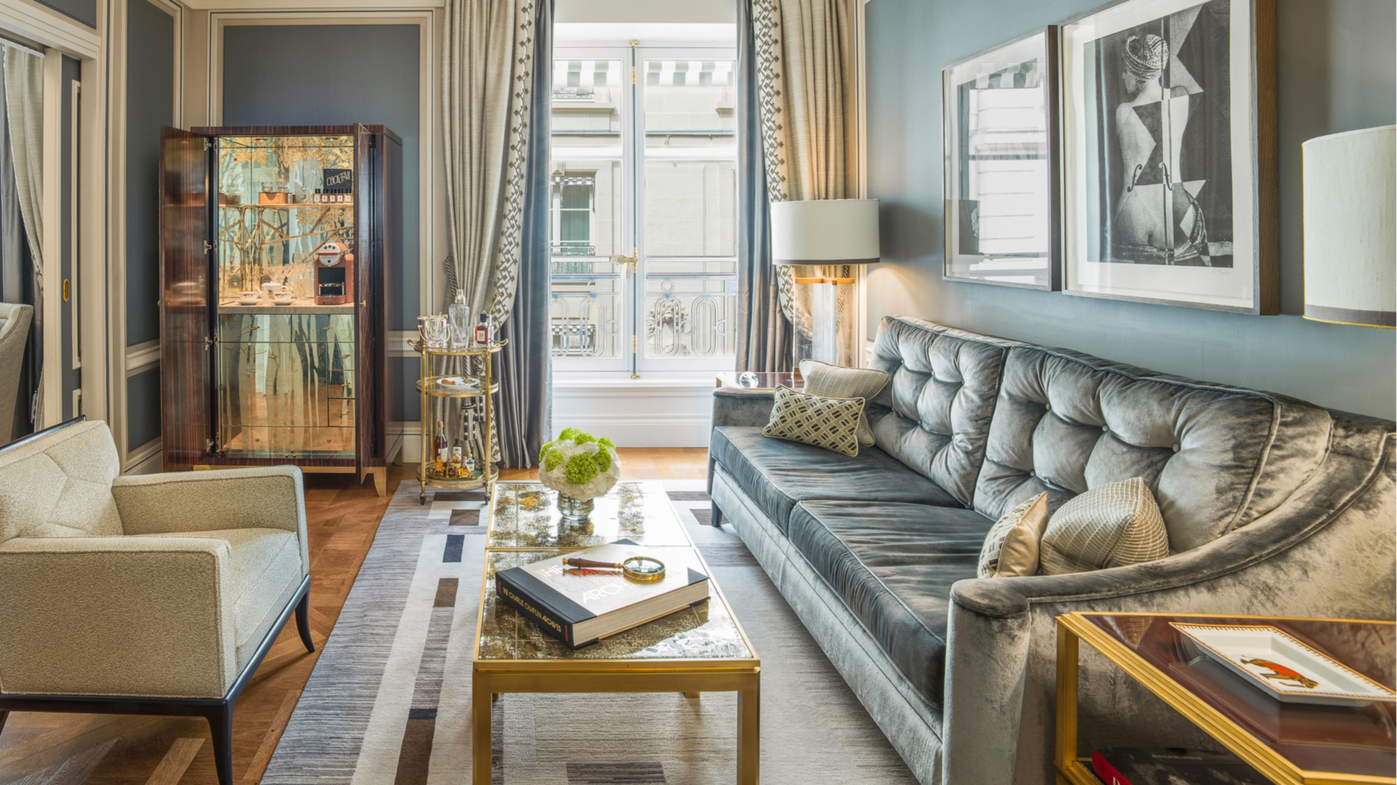 A hotel suite lounge area with a velvet sofa, modern armchairs, a coffee table, a minibar, and large windows.