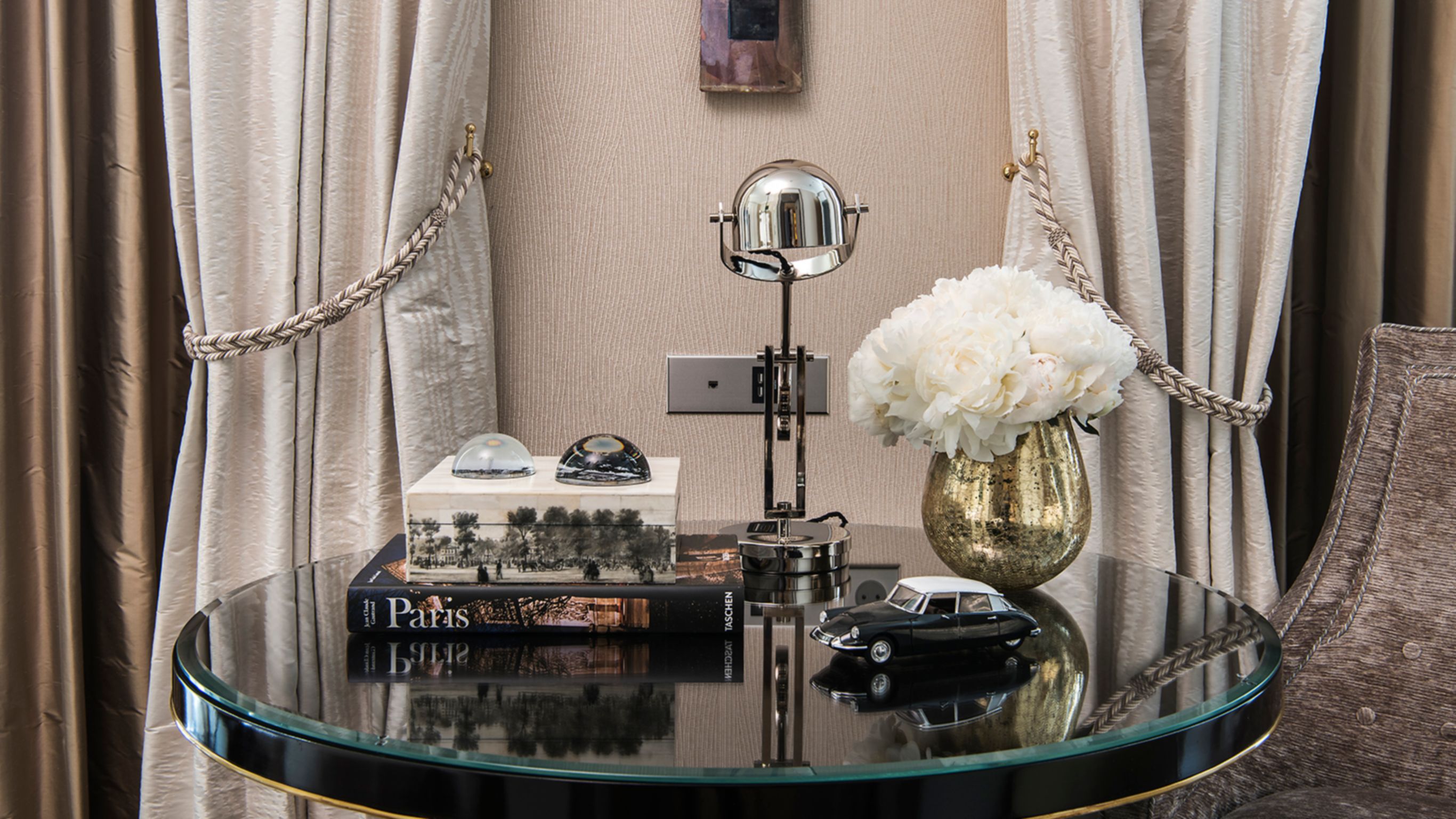 A round glass table holding a vase with white flowers, a model car, a book about Paris, and decorative objects.