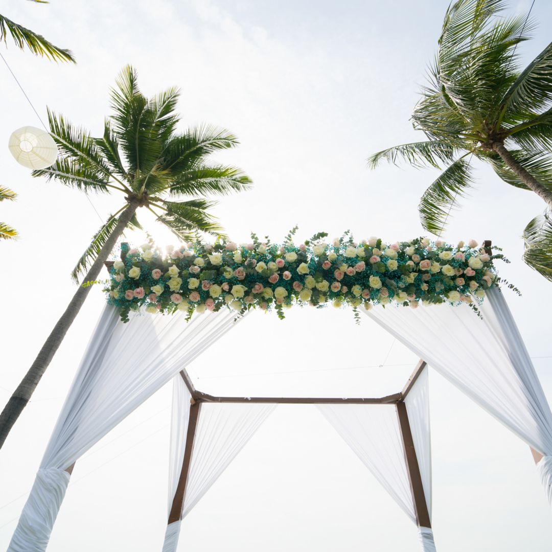 Floral wedding arch under a clear sky, surrounded by palm trees at a Caribbean wedding.