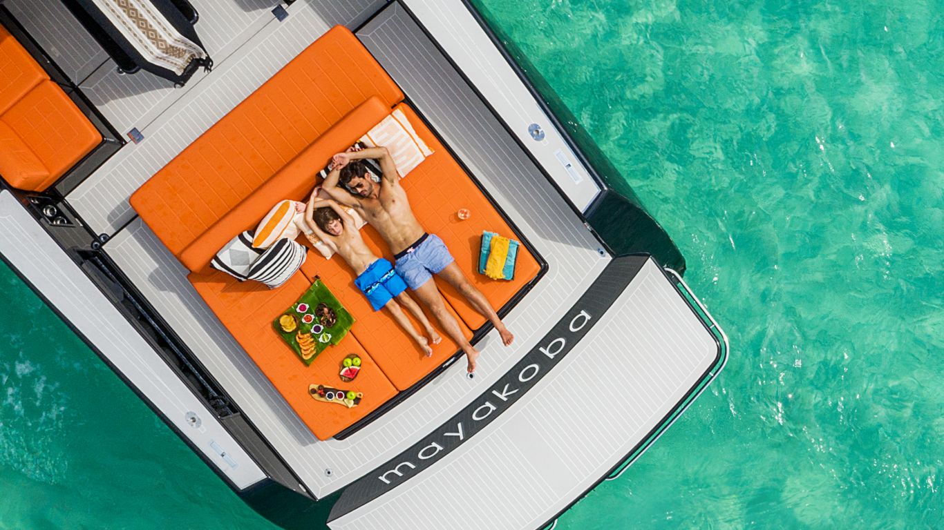 father and son sun bath on back of orange cushioned yacht while mother and sister scuba dive in turquoise ocean