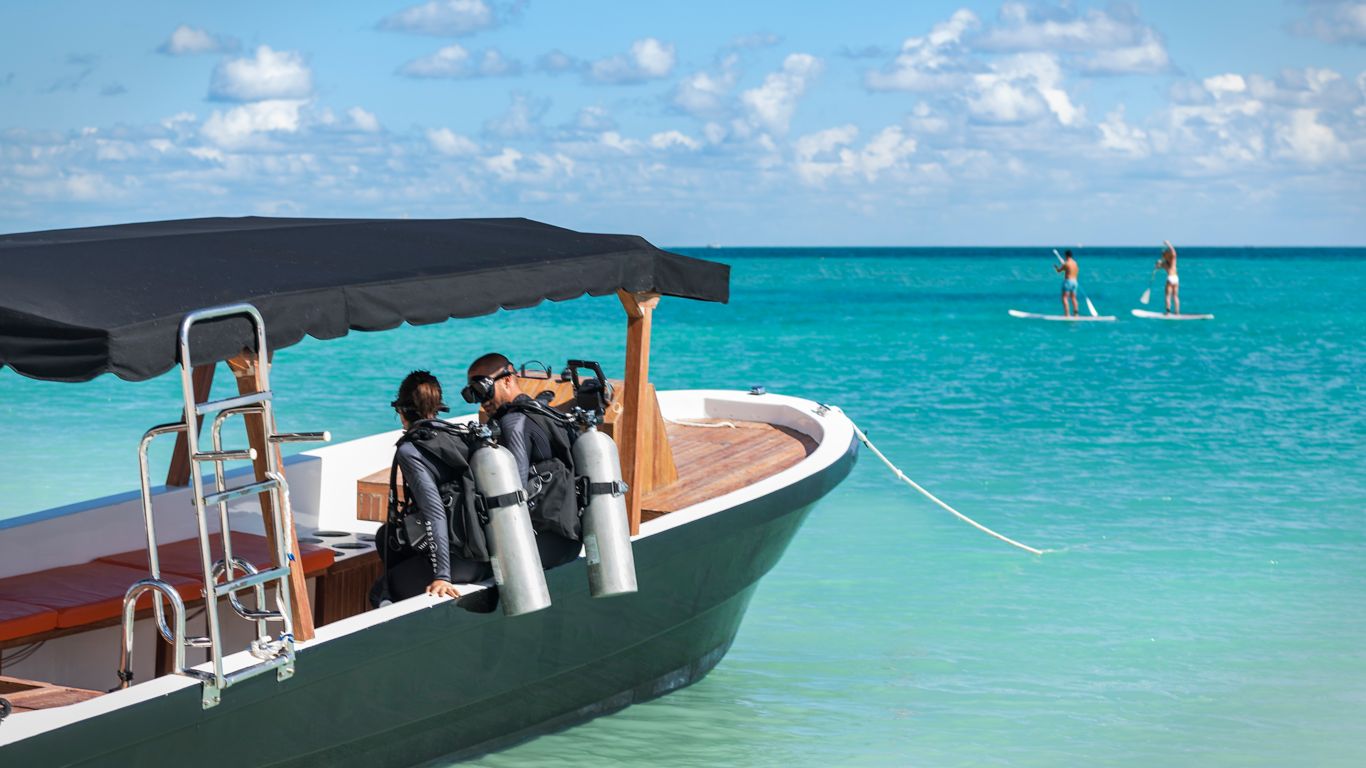two scuba divers with dressed in full equipment sit on anchored boat in turquoise ocea as woman and man paddle board in distance