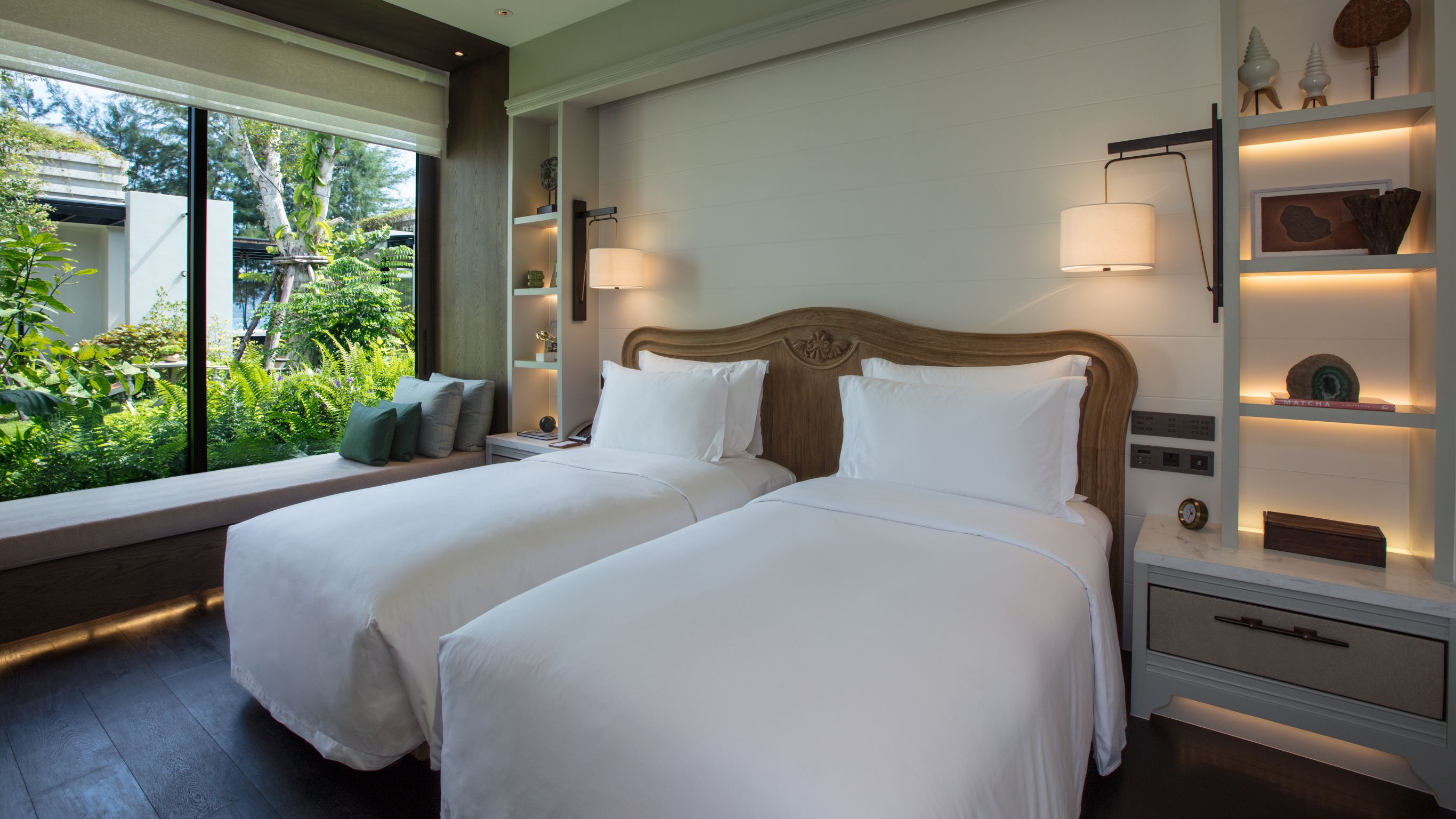 Phuket luxury villa - A hotel room with two single beds, shelves with decorations, dim lights, and windows showing trees outside