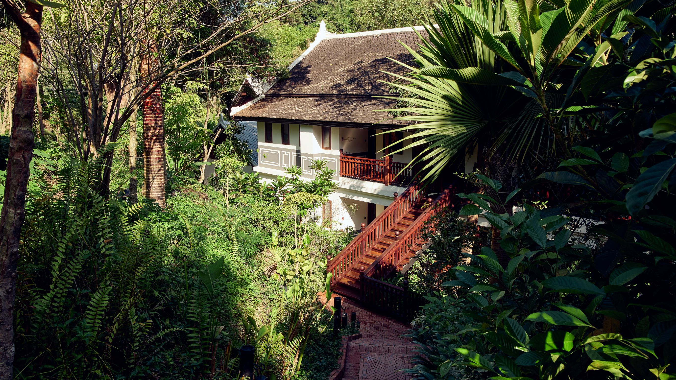 Exterior of a luxury suites in Luang Prabang with white walls, wooden balcony, and stairs amidst lush greenery.