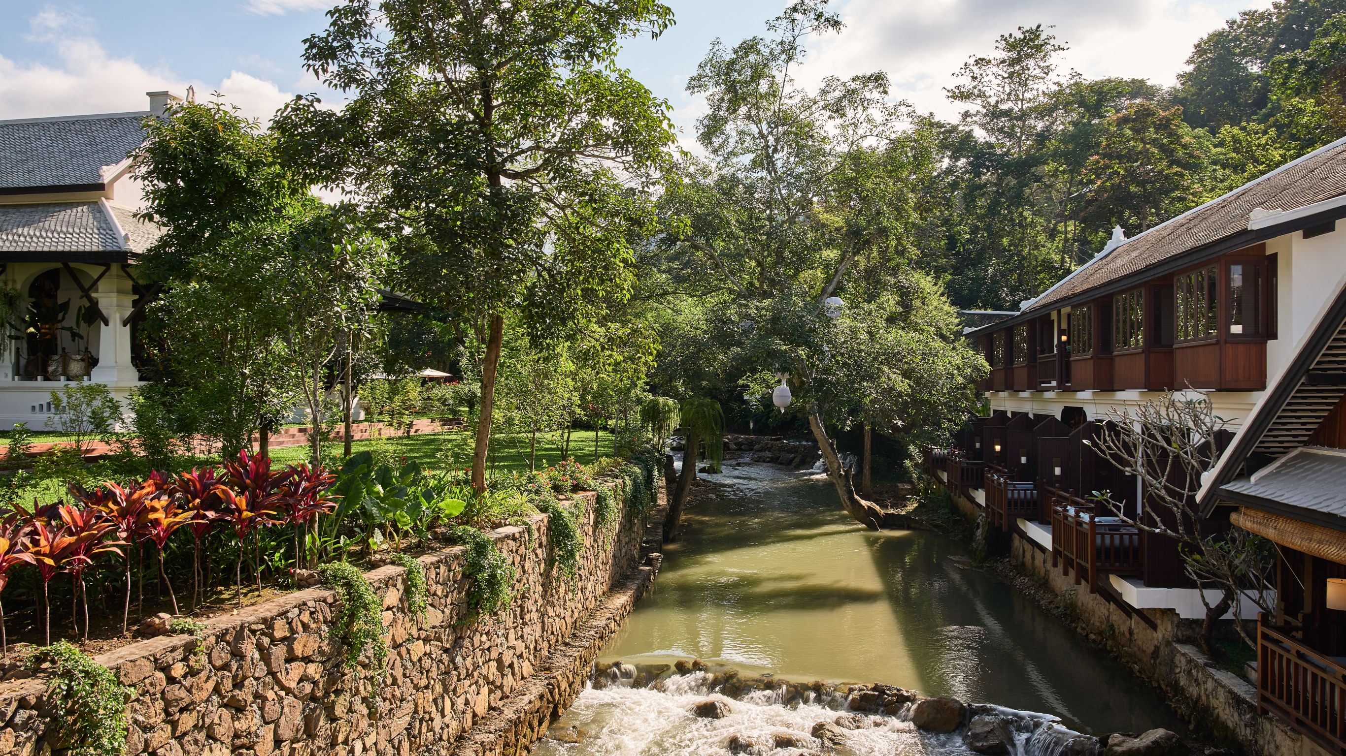 Serene village-like hotel scene with traditional houses, lush greenery, and a flowing river alongside stone walls, in Luang Prabang
