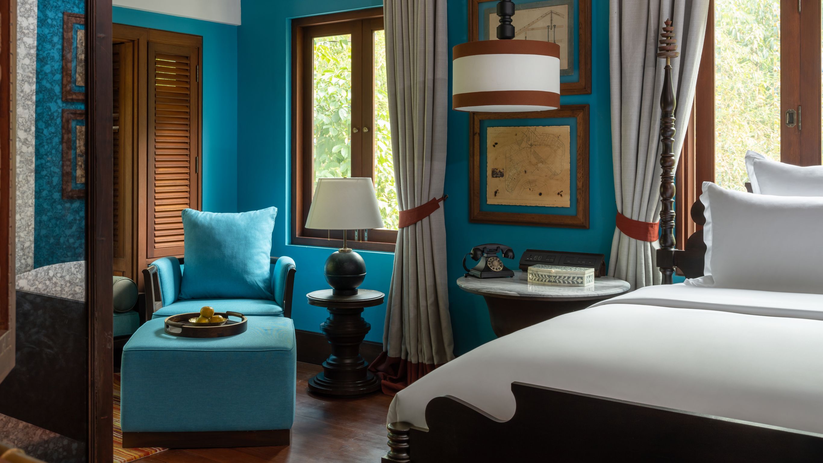 Hotel room in Luang Prabang with blue walls, wooden accents, white bed, blue chair, lamp, and framed art.