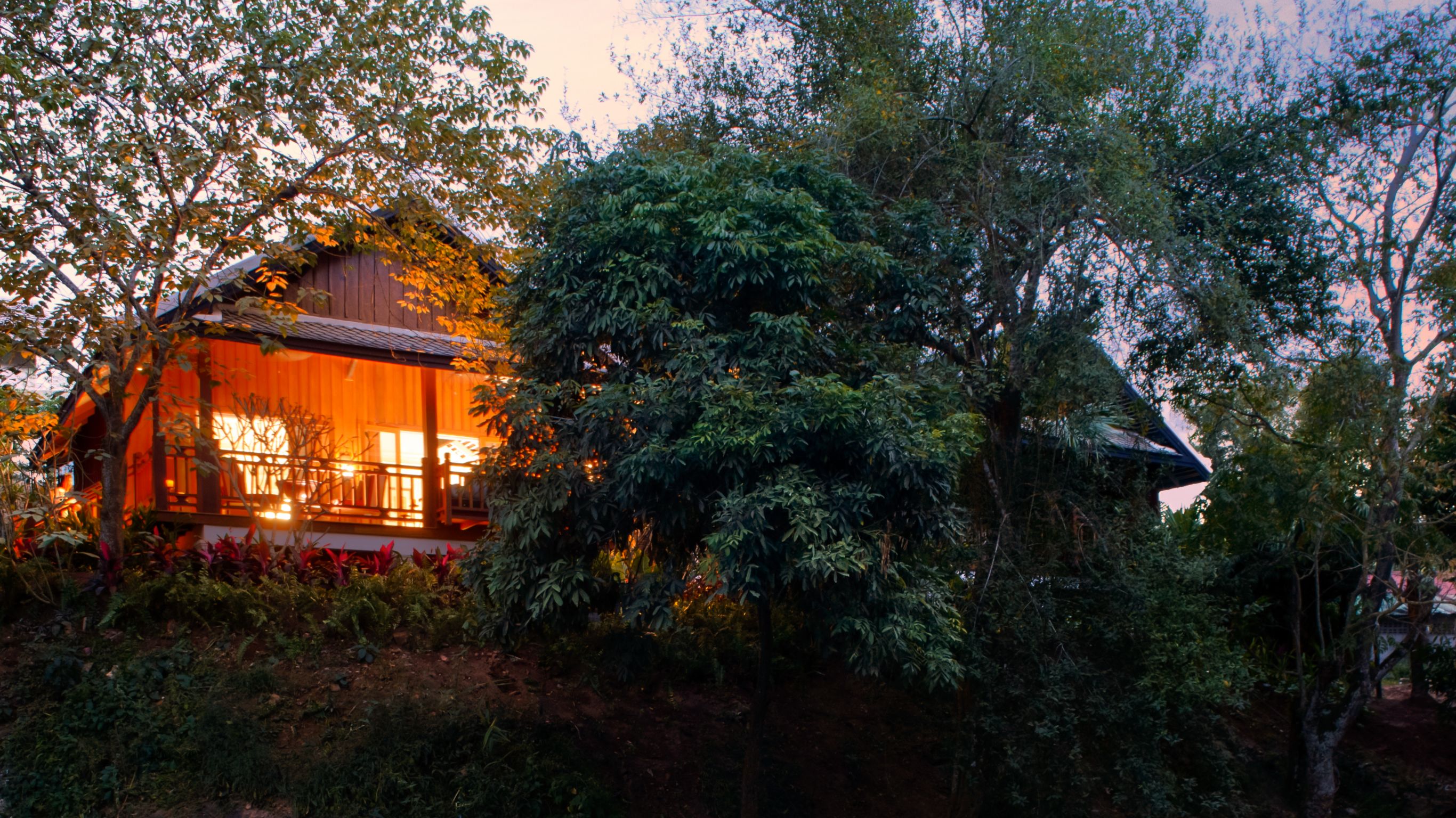 Cozy villa in Luang Prabang with warm lighting, waterfalls, surrounded by lush greenery and trees at dusk.