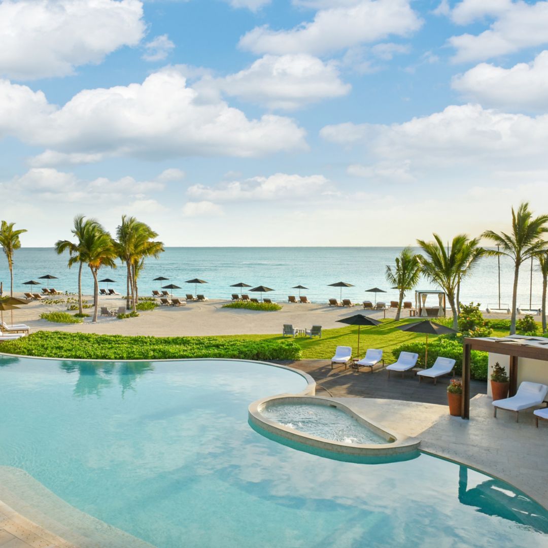 Image Rosewood Mayakoba's Punta Bonita Pool: clear blue water sparlkeling in the sun and teardrop shaped hot tub. Pool is surrounded by pool deck with white lounge chairs and blue umbrellas. Beyond the pool is the beach with palm trees, lounge setups and the caribbean sea.