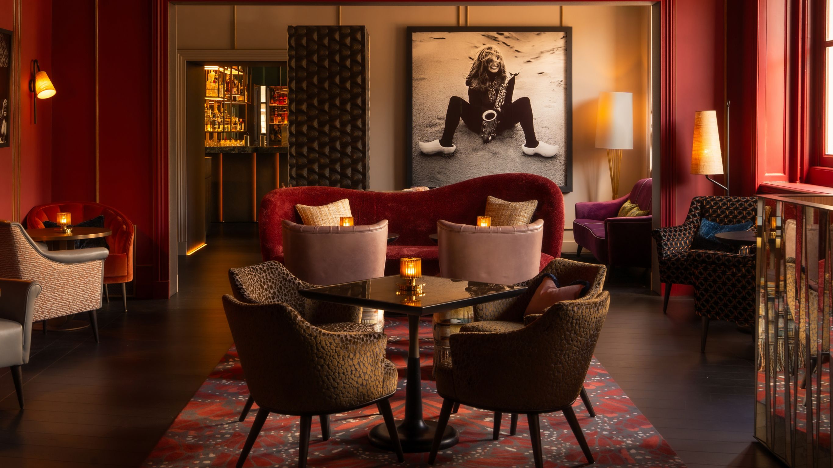 A lounge area decorated in rich reds, featuring patterned chairs, stylish tables, and a black-and-white photo on the wall.