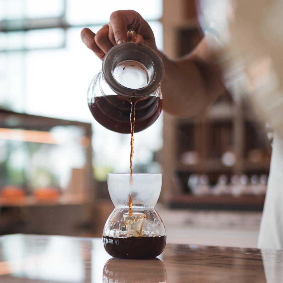 Coffee being poured from a glass container into a glass resting on a wooden table.