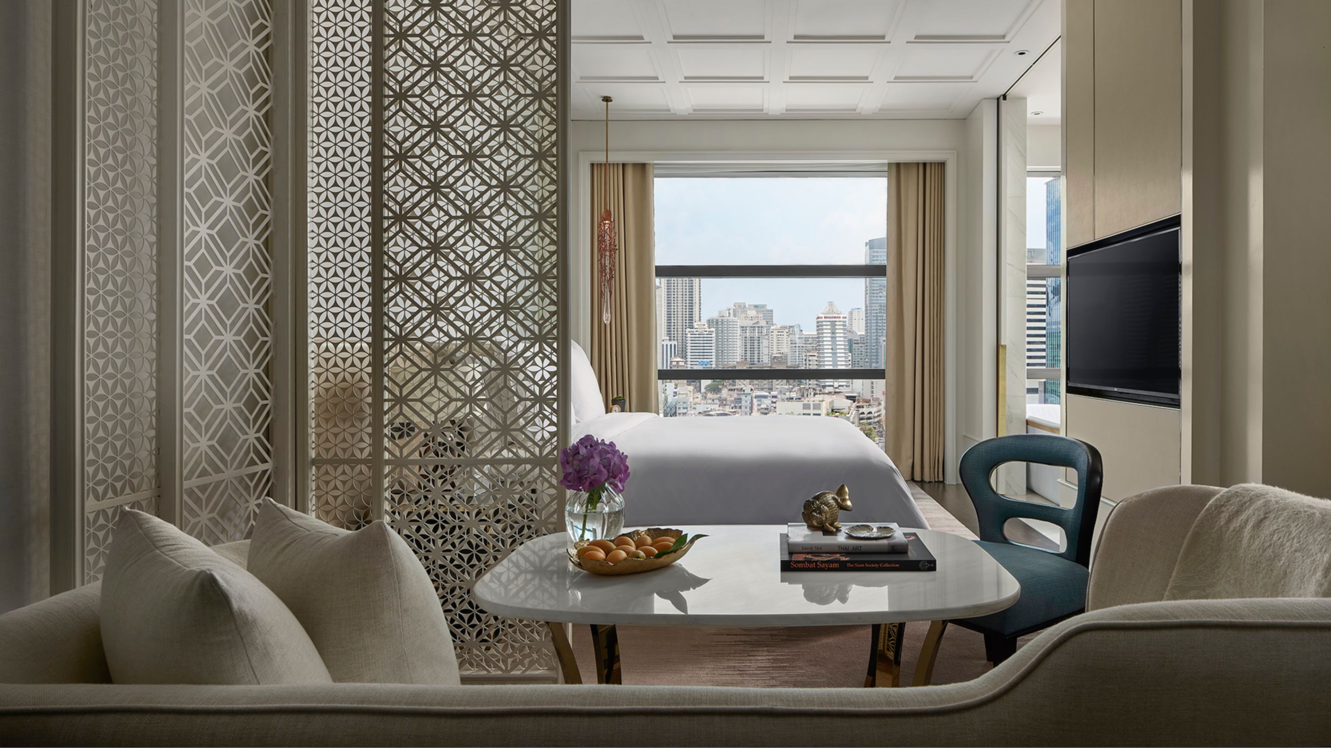 Luxury hotel suite in Bangkok with a large bed facing a TV and a dining area with chairs.