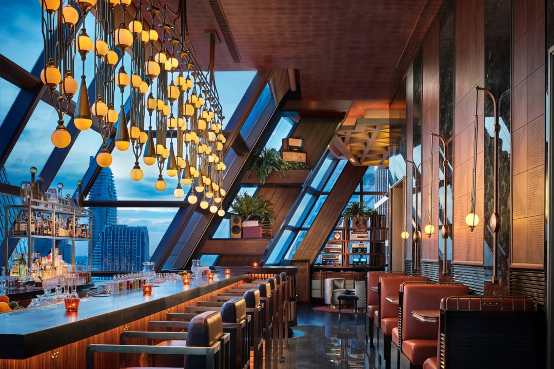 A bar featuring a stunning chandelier, slanted windows, and leather seating, with views of Bangkok.