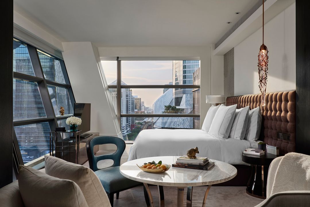 A hotel room in Bangkok with a king-size bed, tufted headboard, a marble table with seating, and a view of the city