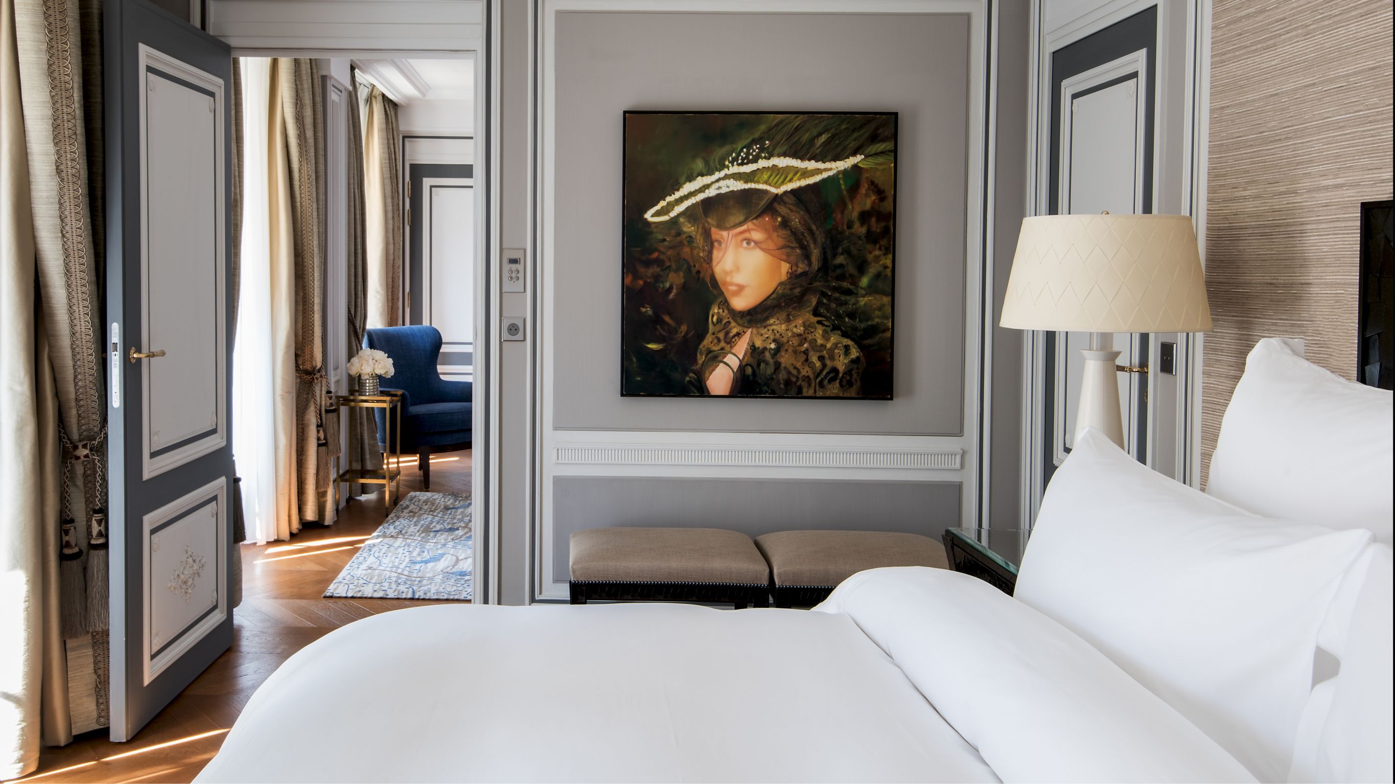 Hotel bedroom with a plush white bed and a portrait of a woman next to an open doorway leading to a sitting area.