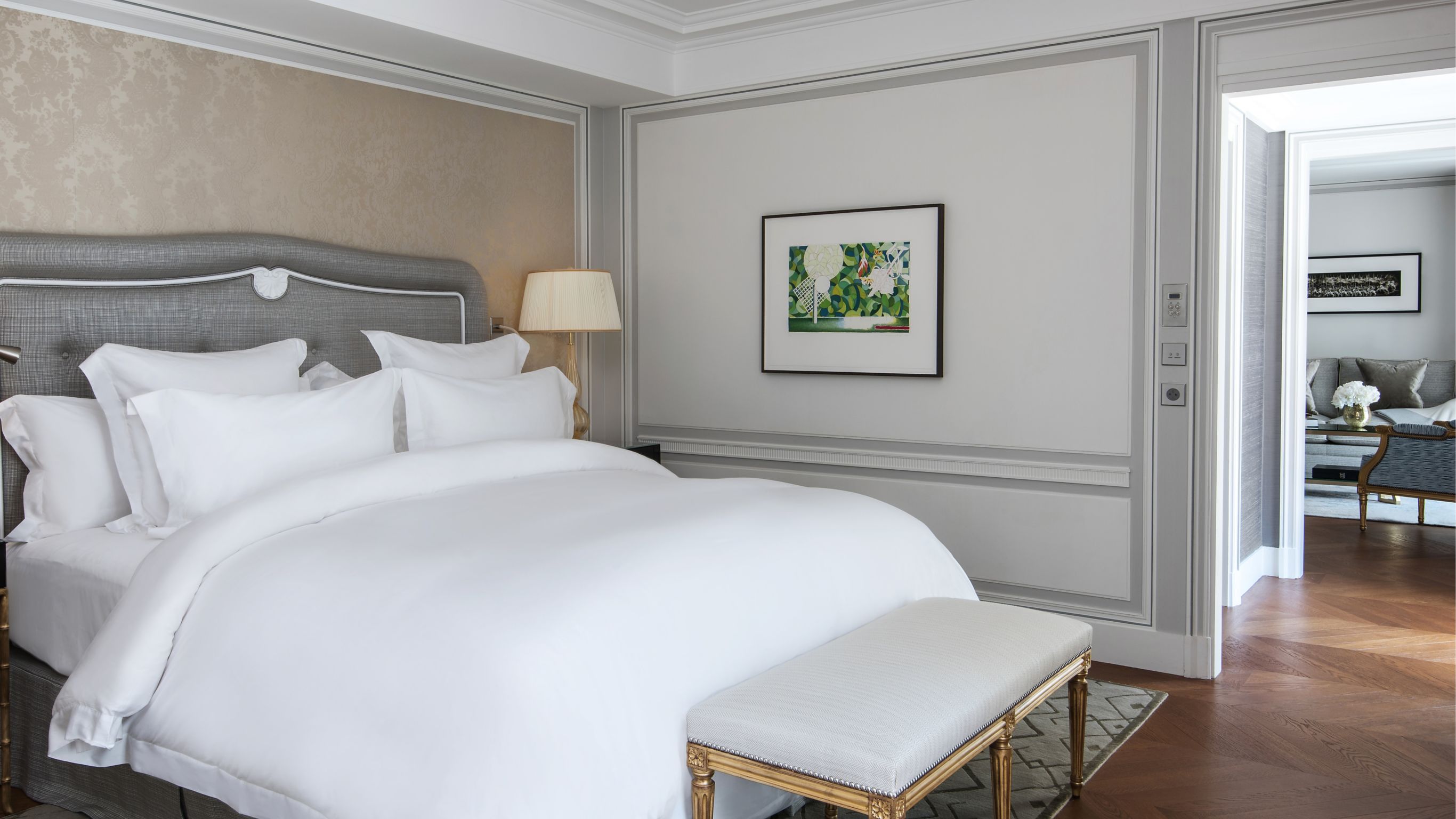 Hotel room with a plush white bed, gold bench and decorations in neutral tones.