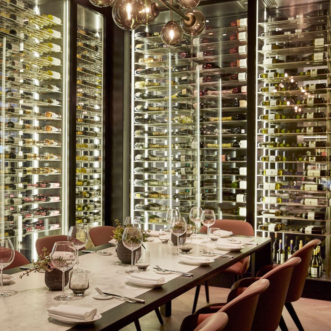 A dining table set with wine glasses and plates, surrounded by chairs, with a backdrop of illuminated wine racks filled with bottle