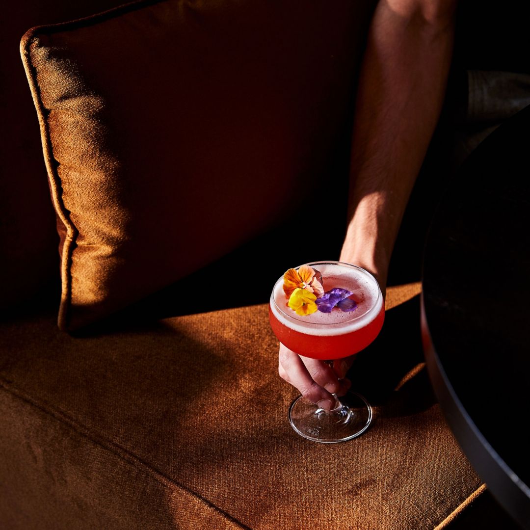 A person holding an orange cocktail resting  on a brown sofa.