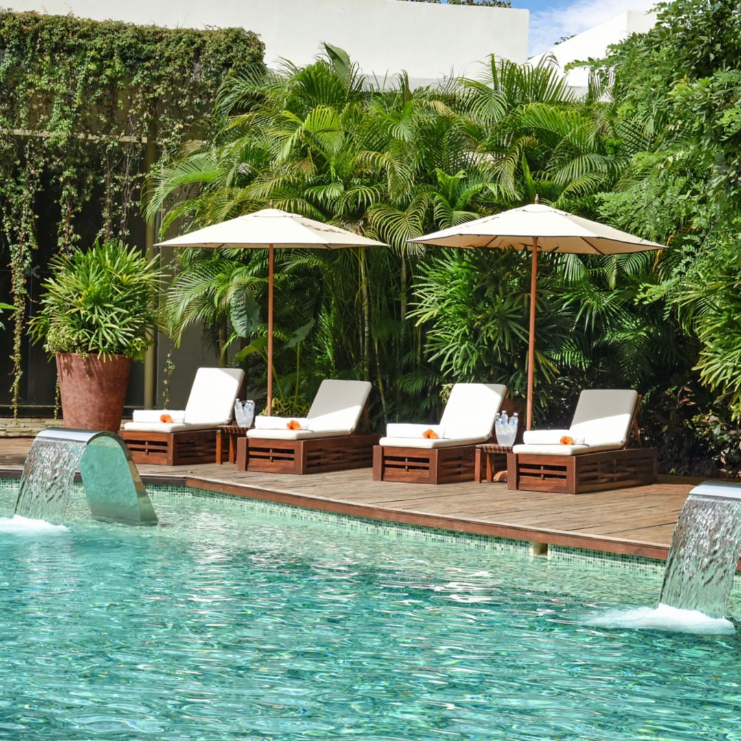 Image Rosewood Mayakoba's Spa Serenity Pool: clear blue water sparkeling in the sun. Pool is surrounded by white lounge chairs, white umbrellas, lush greenery, and planters. There are two fountains at the edge of the pool.