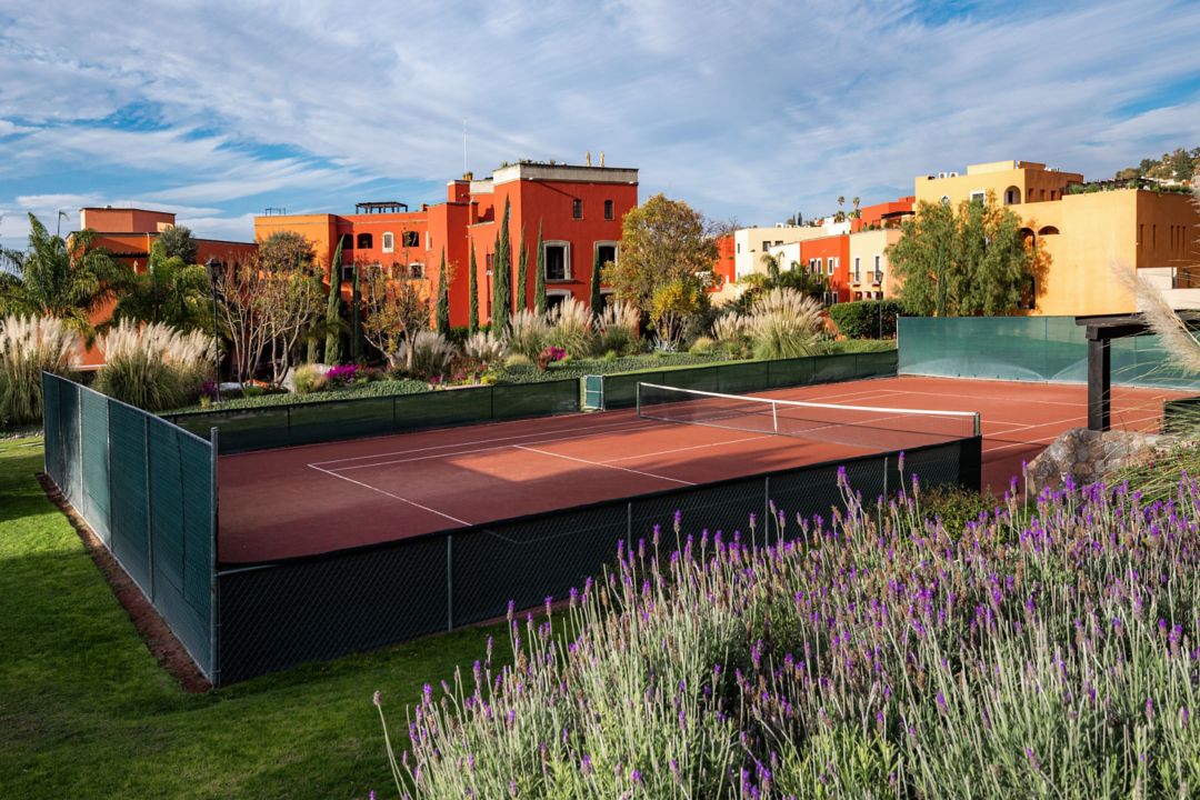 An outdoor tennis court surrounded by vibrant gardens and colorful buildings under a bright sky.