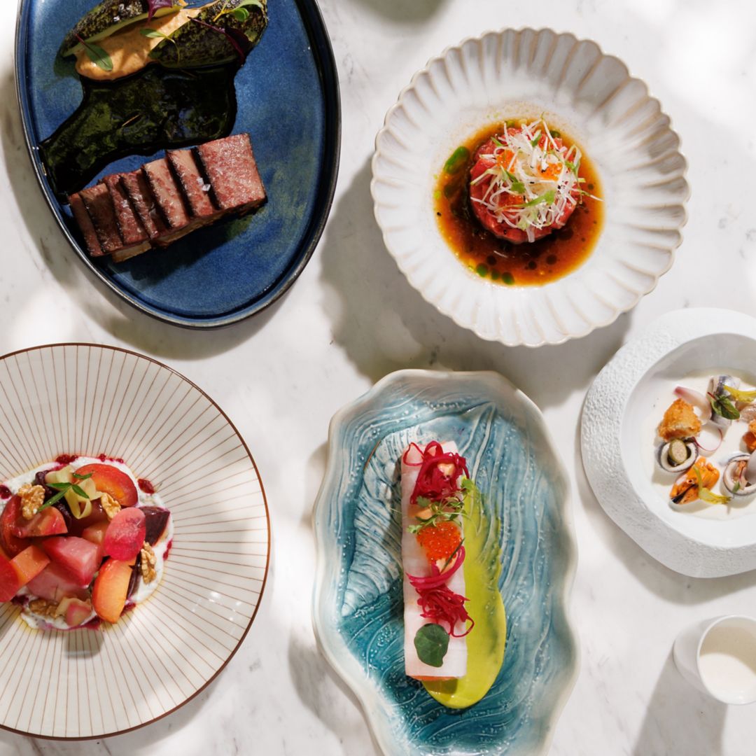 plano cenital de varios platos veraniegos en una terraza sobre mesa de mármol blanca - burrata con piña, ajo blanco,  costilla de vaca,  canelón de salmon