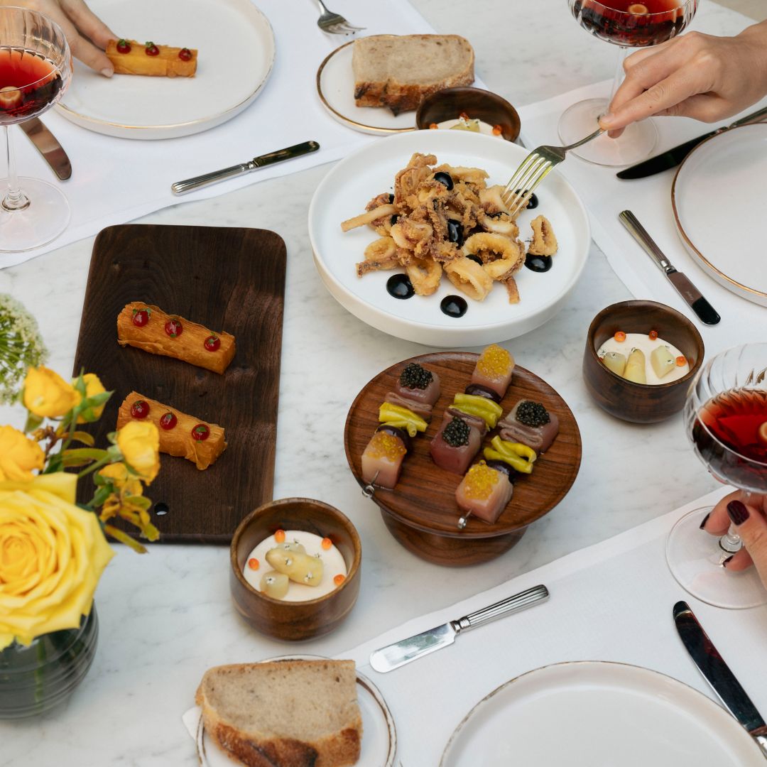 A table with small appetizers, bread, and shared plates of food, surrounded by people