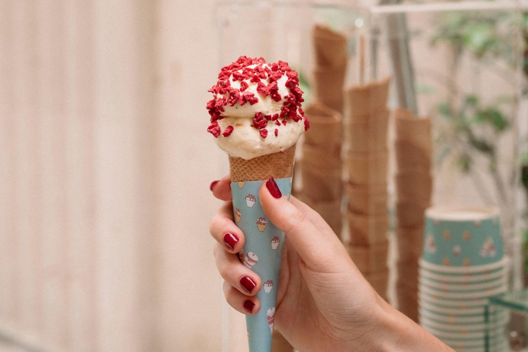A woman's hand holding a vanilla ice cream cone with pink toppings, with an ice cream cart in the background